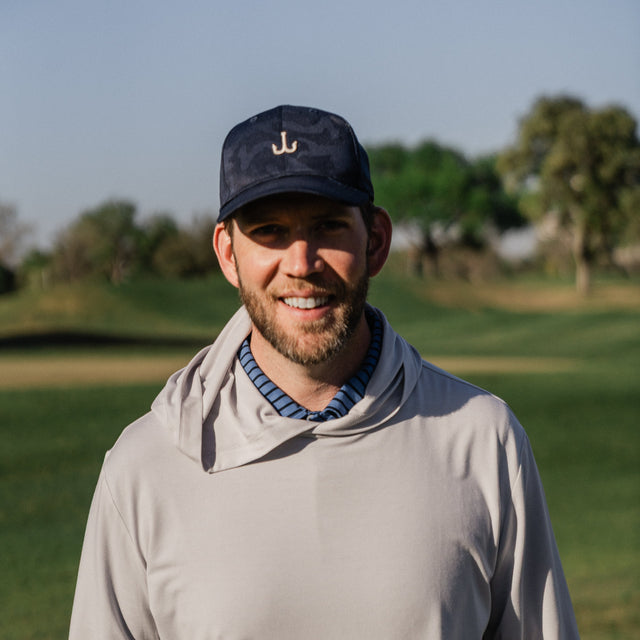 A man wearing a light gray hooded long-sleeve shirt and a navy baseball cap with a white anchor logo, standing on a golf course.