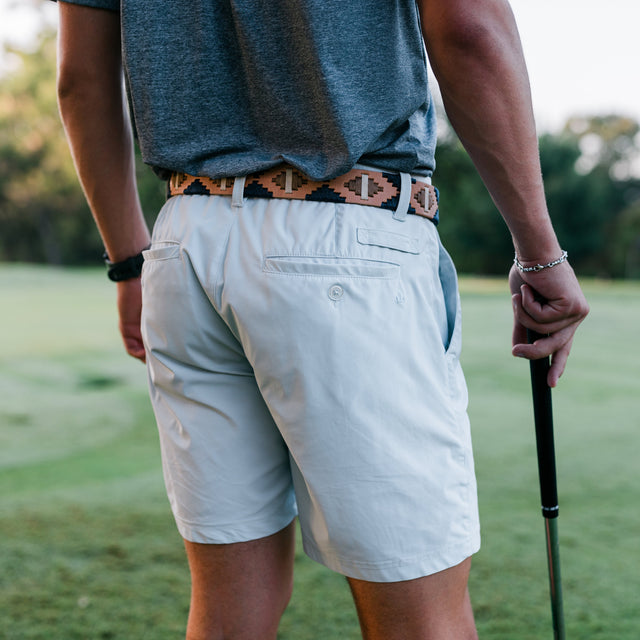 A man on a golf course seen from behind, wearing light beige shorts, a heather gray t-shirt, and a patterned brown belt, holding a golf club.