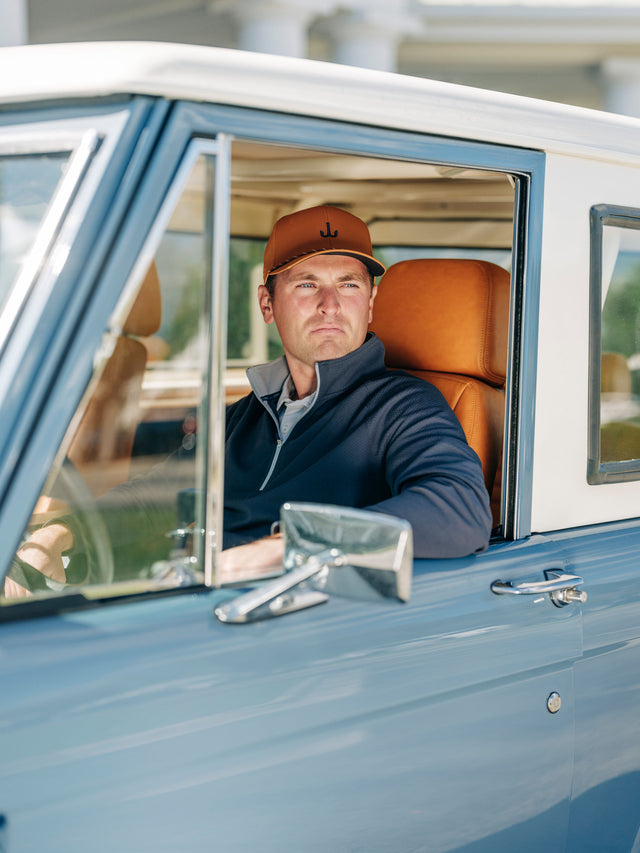 A man wearing a brown baseball cap and navy blue zip-up sweater sits in the driver's seat of a vintage light blue truck with tan leather seats.