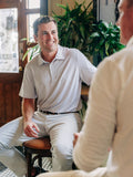 A man in a light gray polo shirt and khaki pants sitting on a stool in a warm, indoor setting with plants in the background, smiling at another person whose back is shown in the foreground.