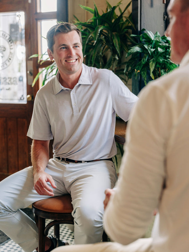 A man in a light gray polo shirt and khaki pants sitting on a stool in a warm, indoor setting with plants in the background, smiling at another person whose back is shown in the foreground.