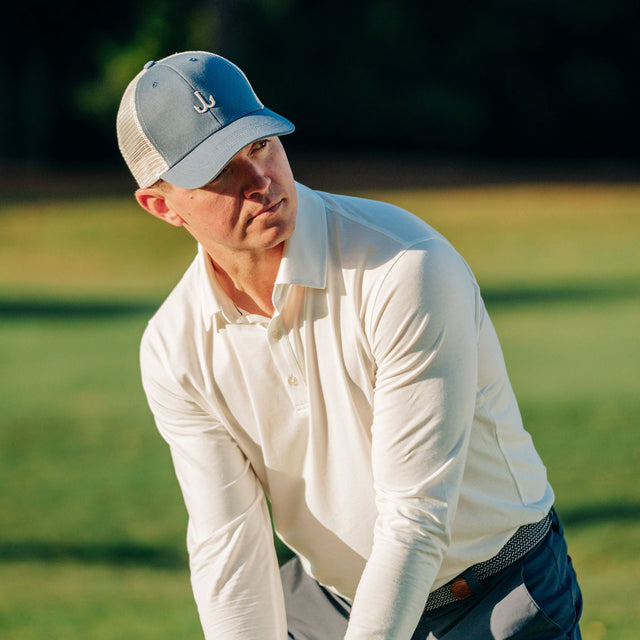 A man on a golf course wearing a light gray and white trucker hat, a long-sleeve white polo shirt, and dark navy pants, captured mid-swing.