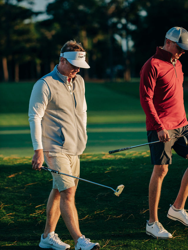 Two men on a golf course at golden hour, one wearing a light gray sleeveless zip vest over a white long-sleeve shirt and khaki shorts, and the other in a red long-sleeve shirt and dark shorts, both holding golf clubs.