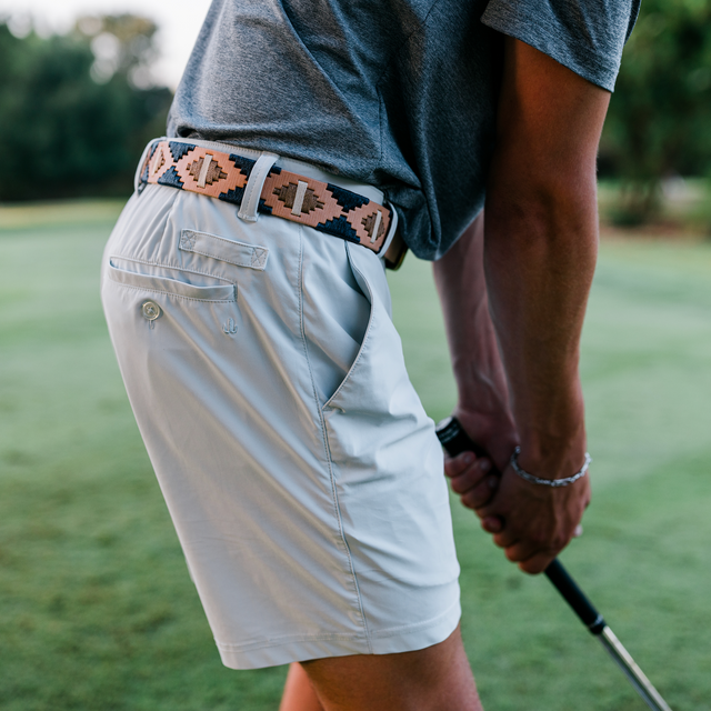A close-up lifestyle shot of a person on a golf course wearing light beige shorts, a gray t-shirt, and a patterned belt with a prominent 'D' logo, holding a golf club.
