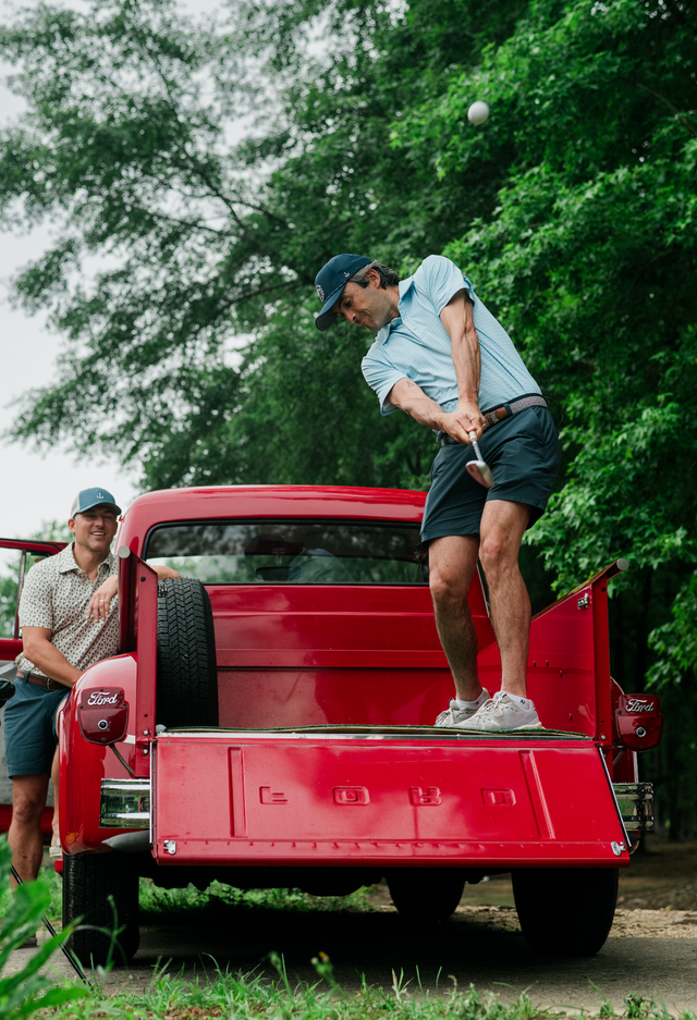 Man playing golf on a red truck bed with another person watching.