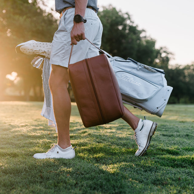 A person walking on a grassy golf course at sunset, carrying a brown leather golf bag and a light gray golf bag over their shoulder.